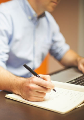 Imagem de uma pessoa escrevendo com caneta em um caderno e ao mesmo tempo usando um notebook